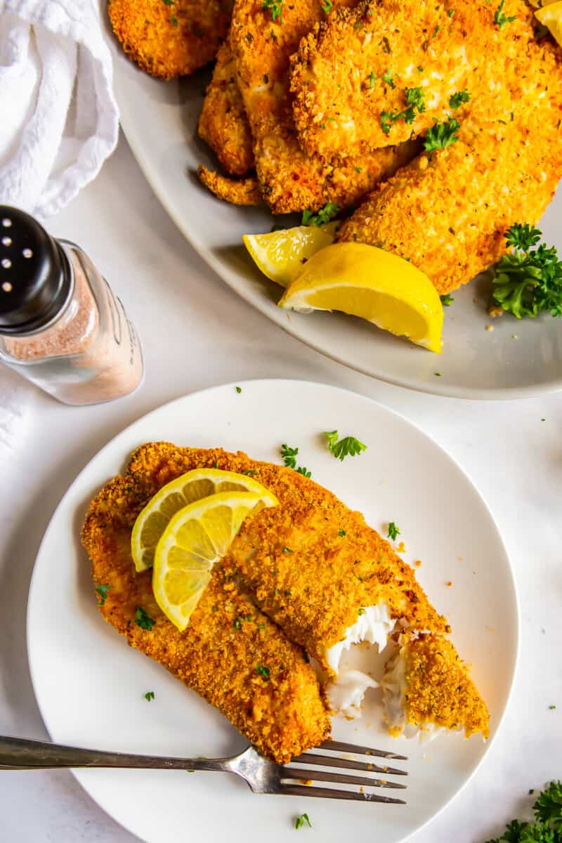 overhead image of bite out of breaded tilapia filet topped with lemon slices on a white plate with a fork