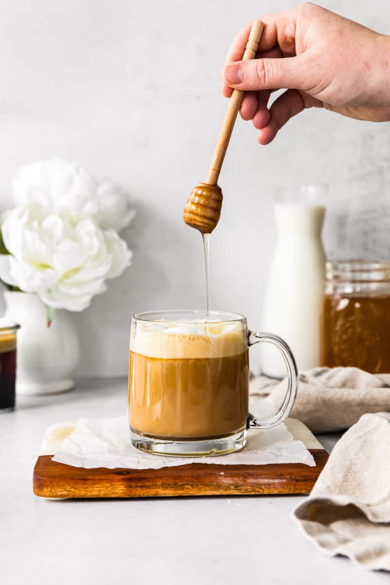 hand drizzling honey into honey almond milk flat white in a clear glass mug