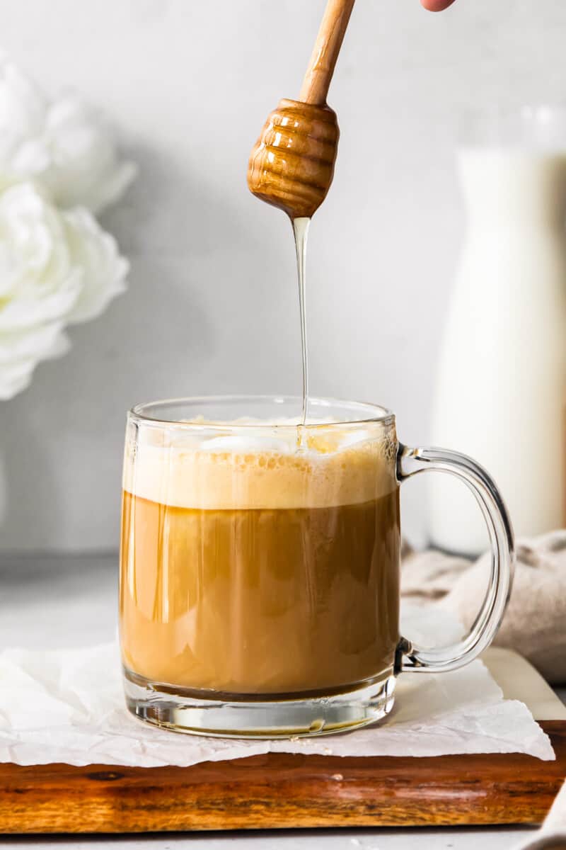 drizzling honey into honey almond milk flat white in a clear glass mug