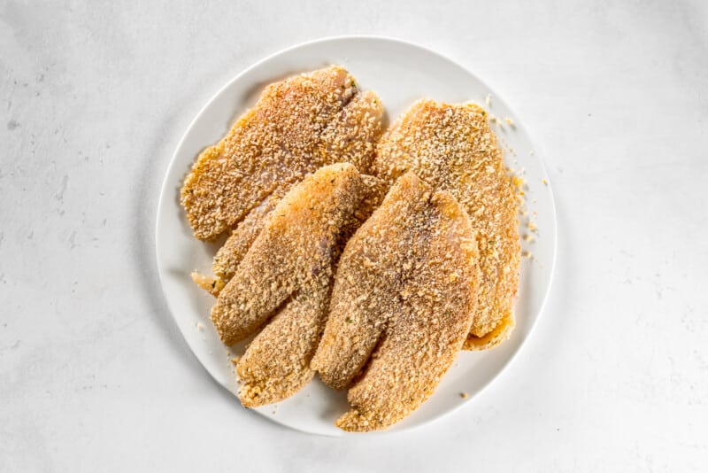 4 breaded tilapia filets on a white plate before cooking