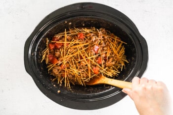 hand stirring spaghetti noodles and meat sauce together in crock pot
