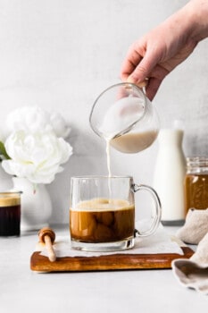 hand pouring almond milk into espresso in a clear mug