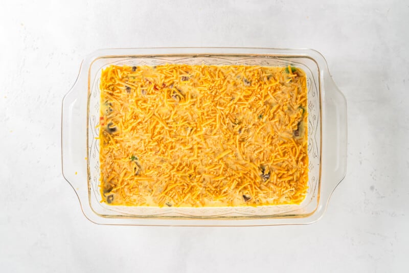 sausage egg breakfast casserole mixture topped with shredded cheese in a glass casserole pan before baking
