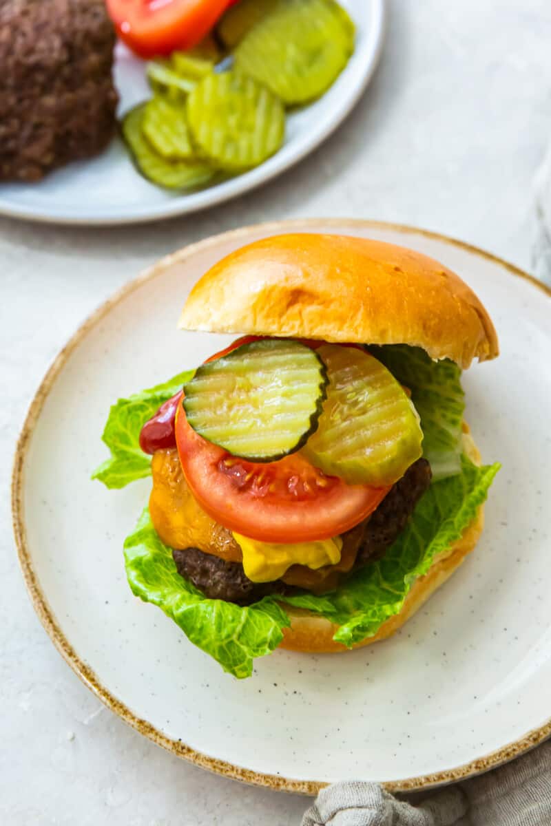 cheeseburger with a bun on a white plate topped with lettuce, cheese, ketchup, tomato slice, and pickles