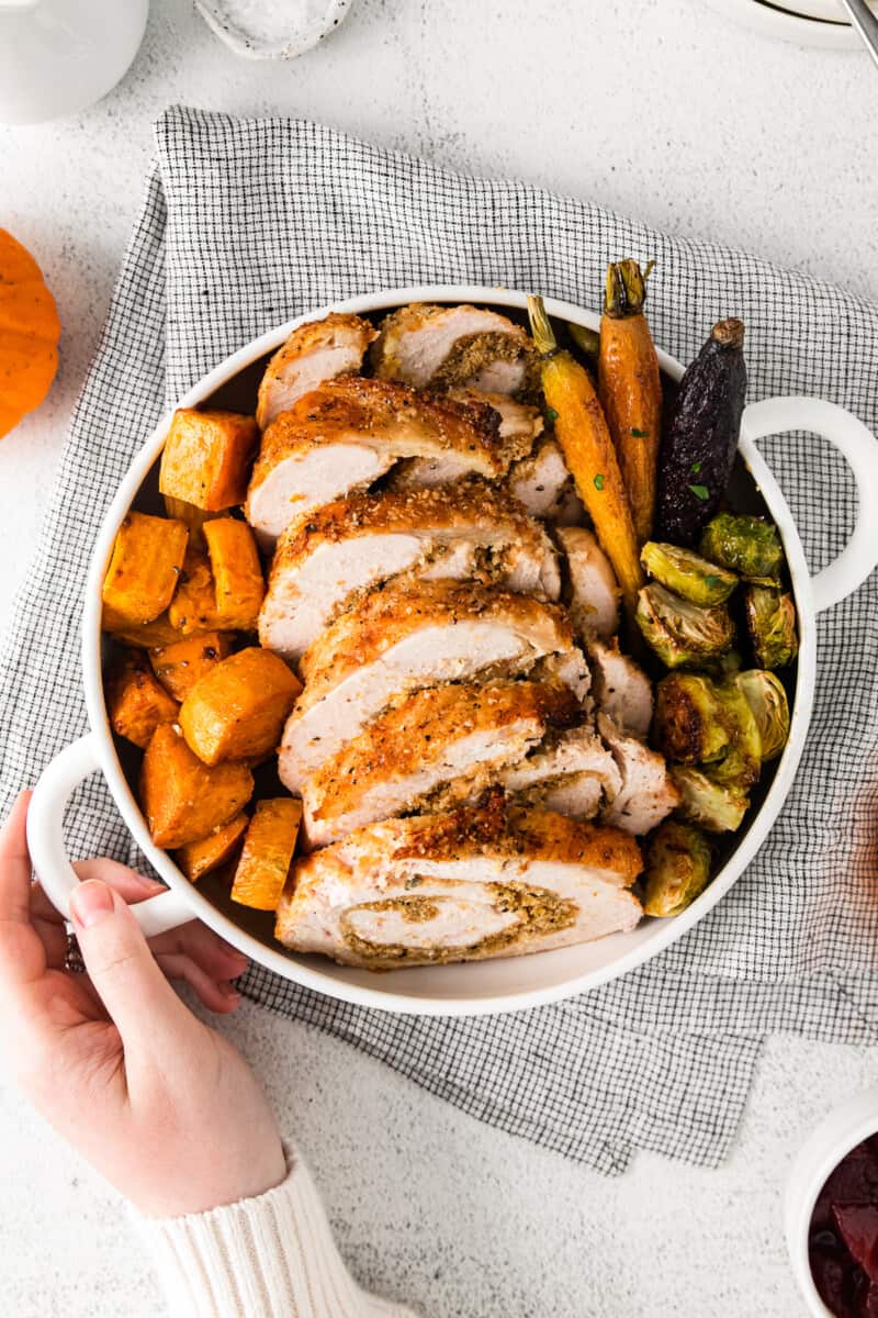 hand holding white plate of roasted sweet potatoes, carrots, and Brussels sprouts and sliced turkey roulade