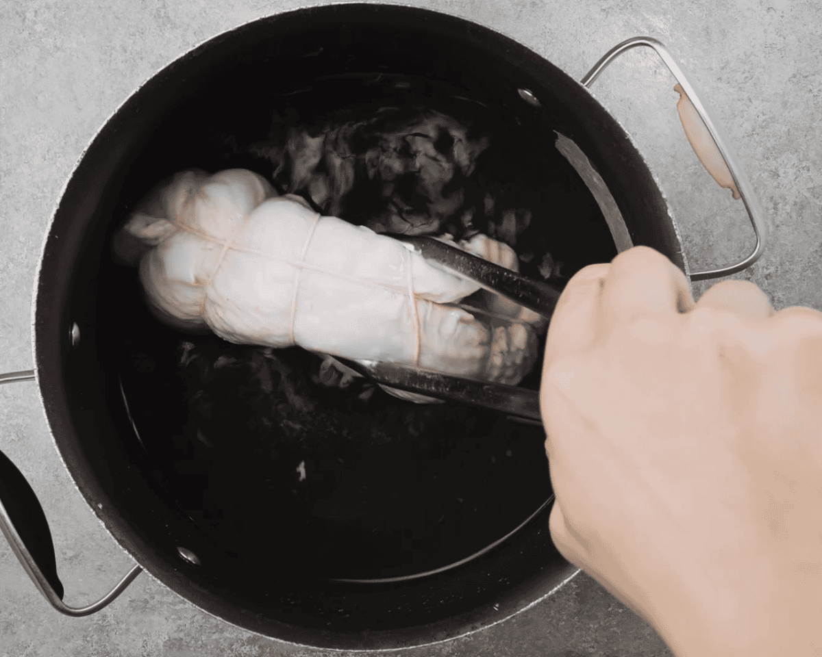 Raw turkey breast tied with kitchen twine being lowered into a large stock pot of water using tongs.