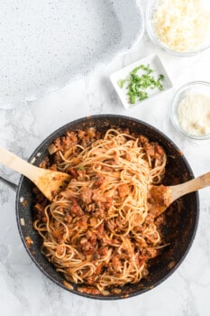 cooked spaghetti noodles coated with marinara meat sauce in a skillet with 2 wood spoons