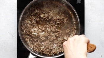 ground beef, onion, and garlic cooking in a skillet with a hand holding a wooden spoon