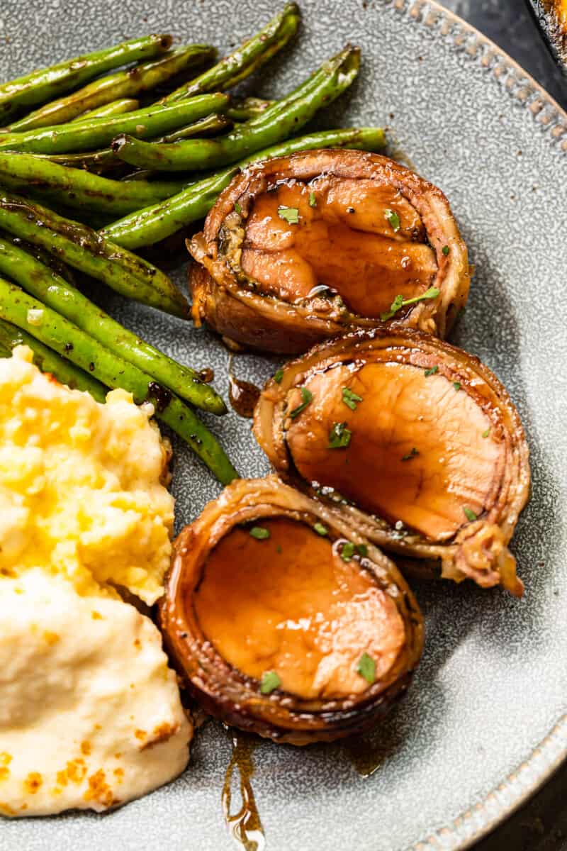 slices of bacon wrapped pork tenderloin on a plate with mashed potatoes and vegetables