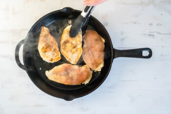 hand using a tong flipping a chicken breast over in a skillet