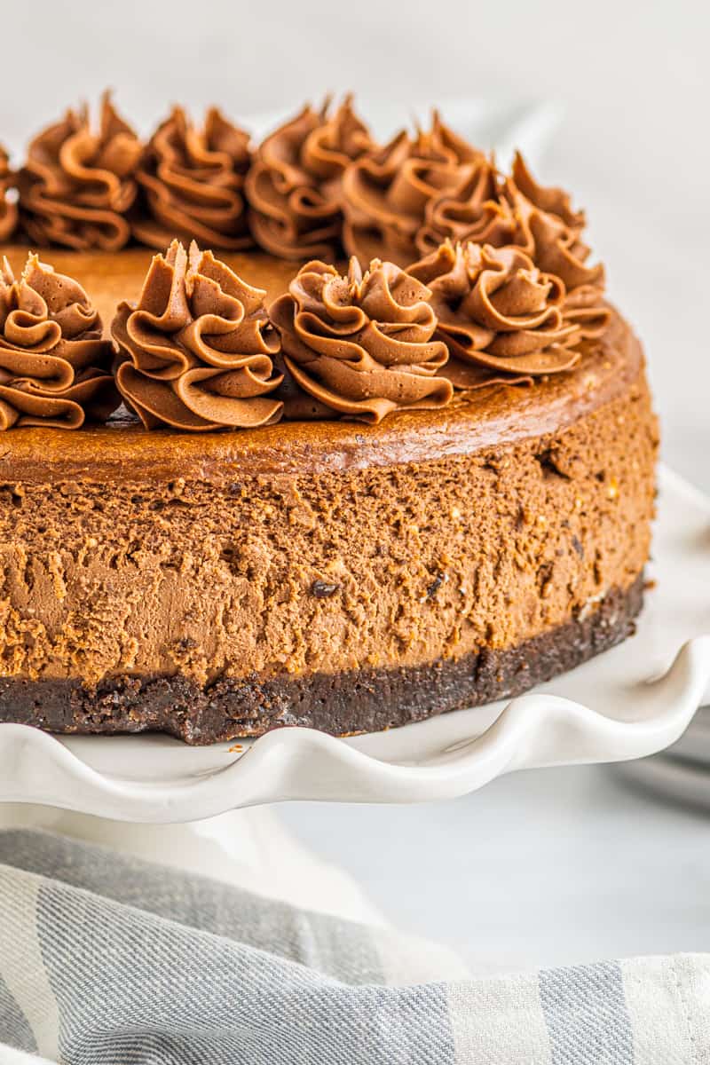 chocolate cheesecake topped with swirls of whipped chocolate frosting on a white cake stand