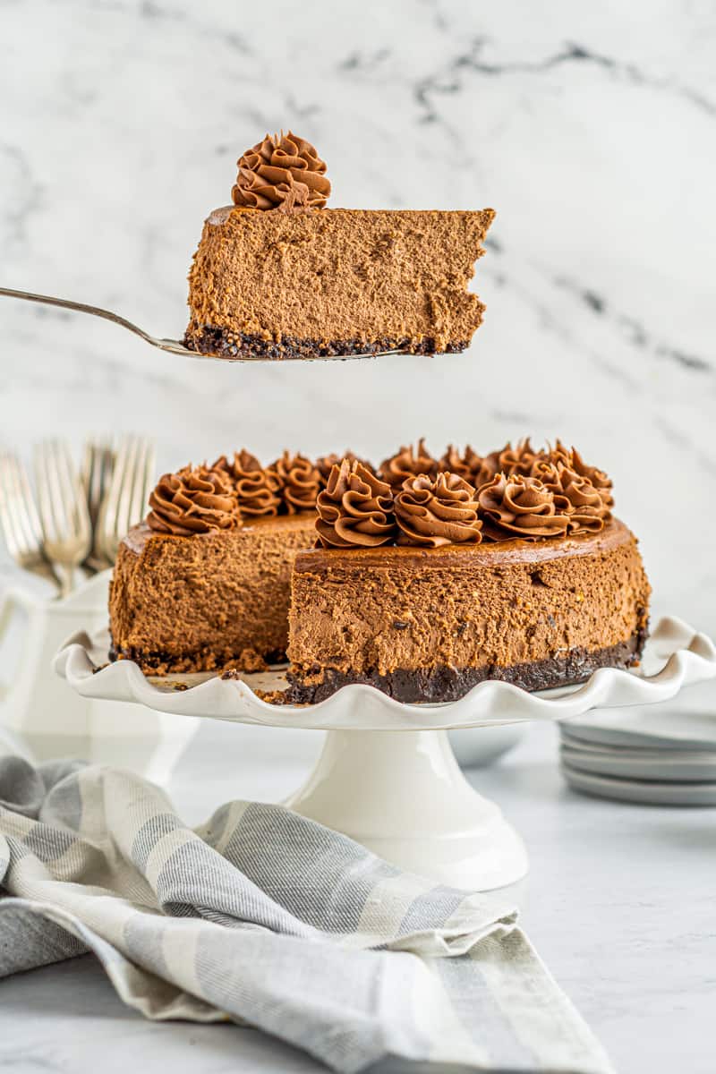 slice of chocolate cheesecake on a dessert server being removed from the full chocolate cheesecake on a white cake stand