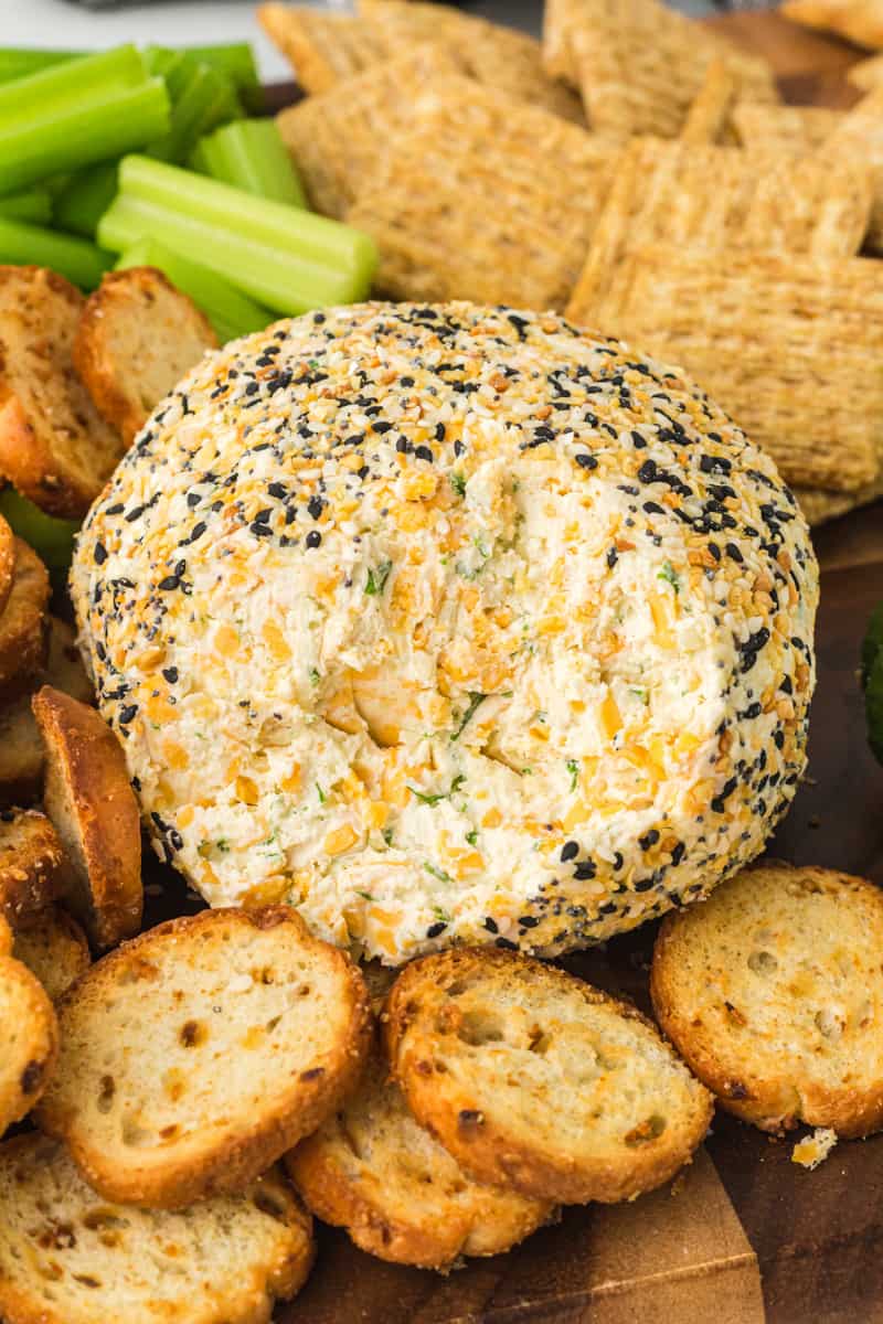 everything bagel cheeseball on a serving board with crackers, sliced cucumbers, and celery sticks with some of the cheeseball gone