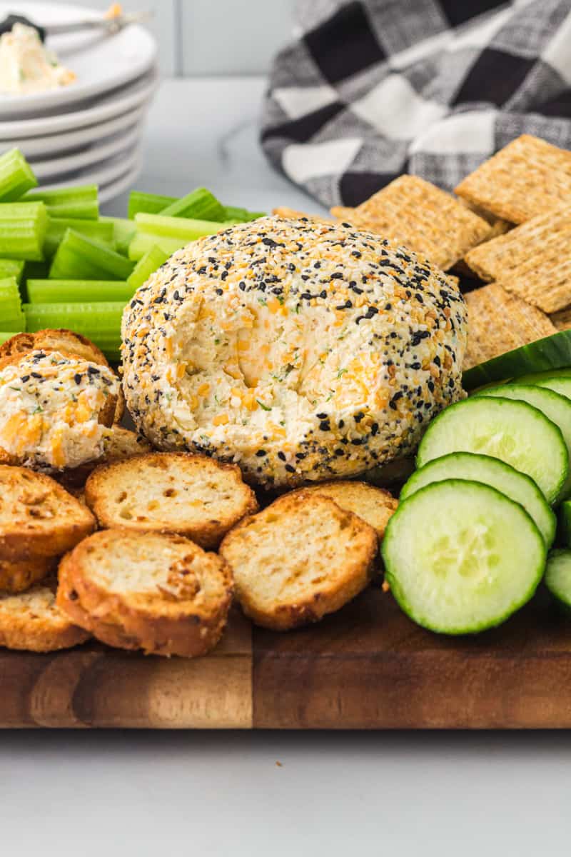 everything bagel cheeseball on a serving board with crackers, sliced cucumbers, and celery sticks with some of the cheeseball gone