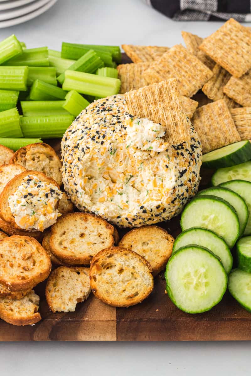 everything bagel cheeseball on a serving board with crackers, sliced cucumbers, and celery sticks with some of the cheeseball gone