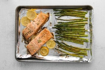 two baked salmon fillets with lemon slices and asparagus on a baking sheet.