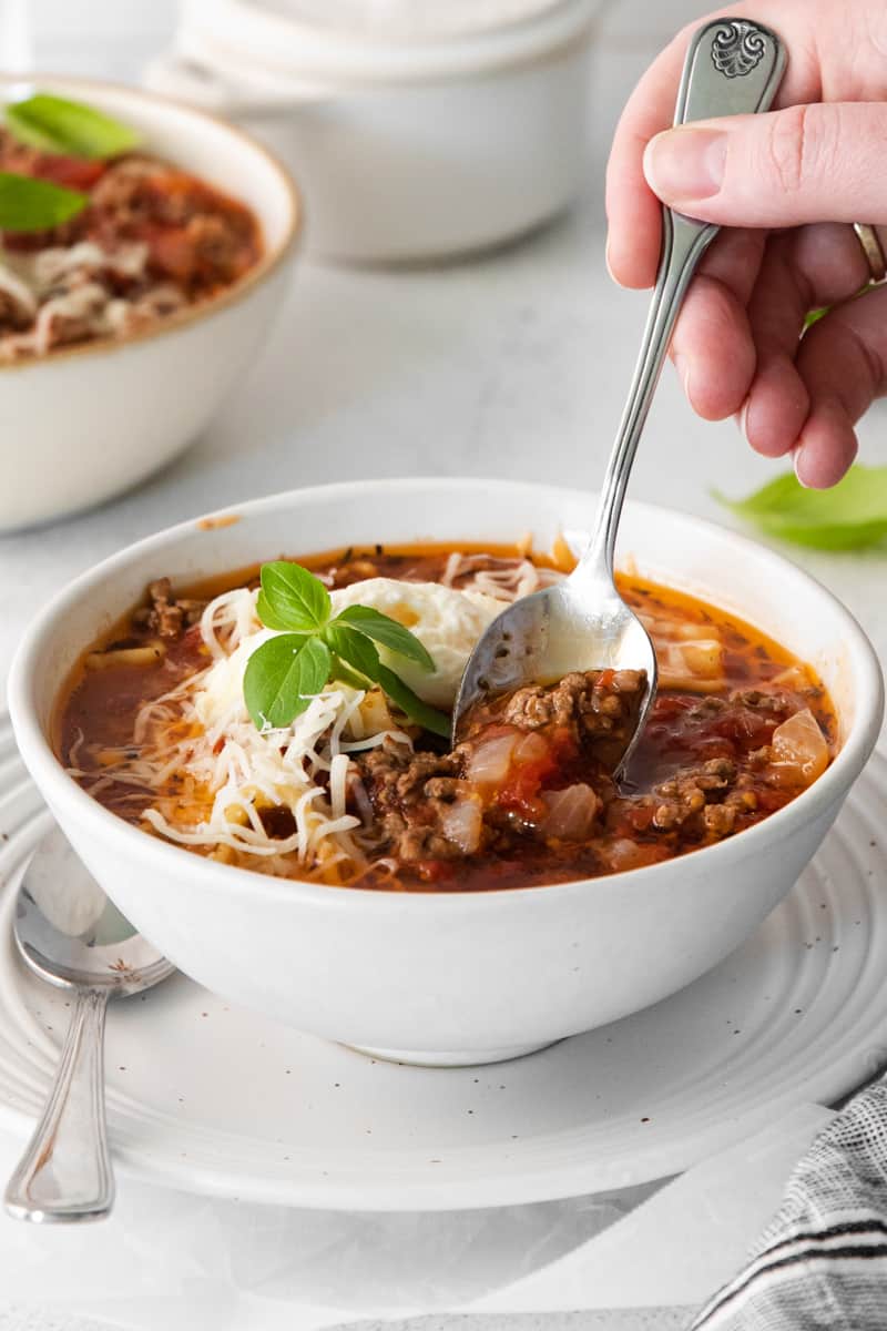 hand with a spoon in a bowl of lasagna soup topped with ricotta cheese, parmesan cheese, and basil