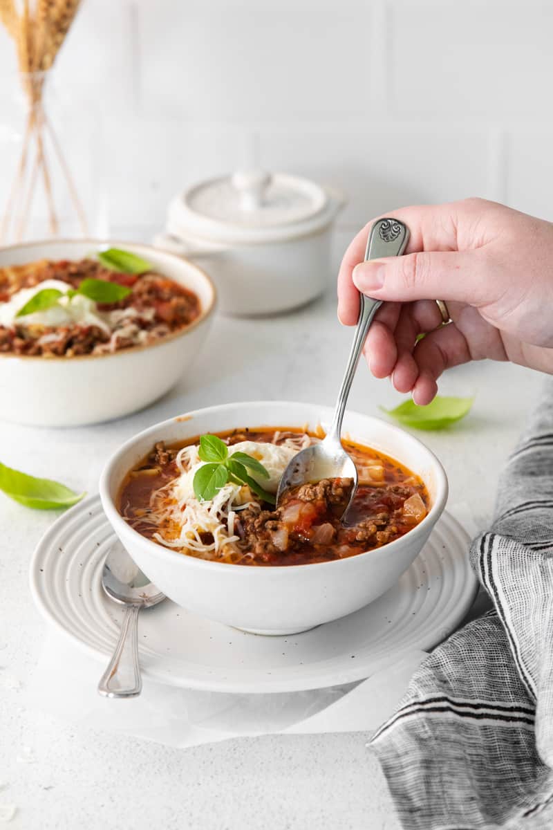 hand with a spoon in a bowl of lasagna soup topped with ricotta cheese, parmesan cheese, and basil