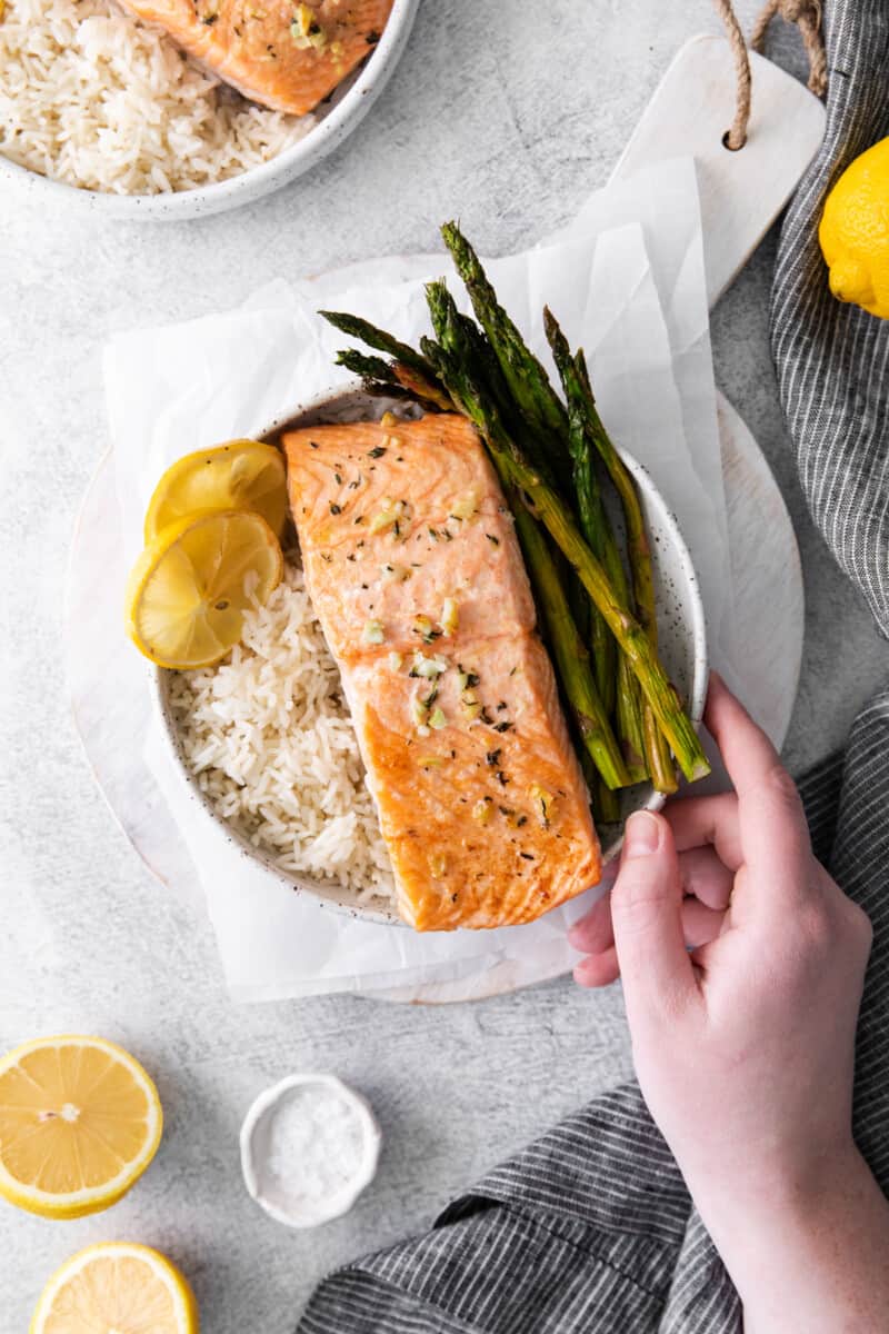 a hand grabbing a white bowl filled with sheet pan lemon salmon on a bed of rice with asparagus.