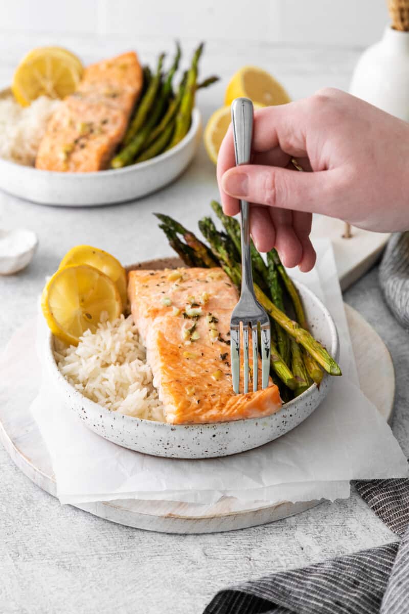 a forkful of sheet pan lemon salmon in front of a bowl of sheet pan lemon salmon on a bed of rice with asparagus in a white bowl.
