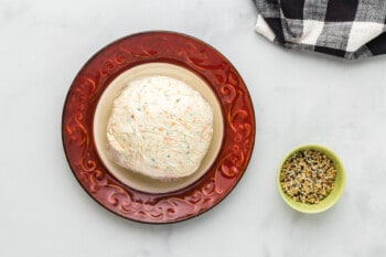 everything bagel cheeseball wrapped in plastic wrap on a red and cream plate