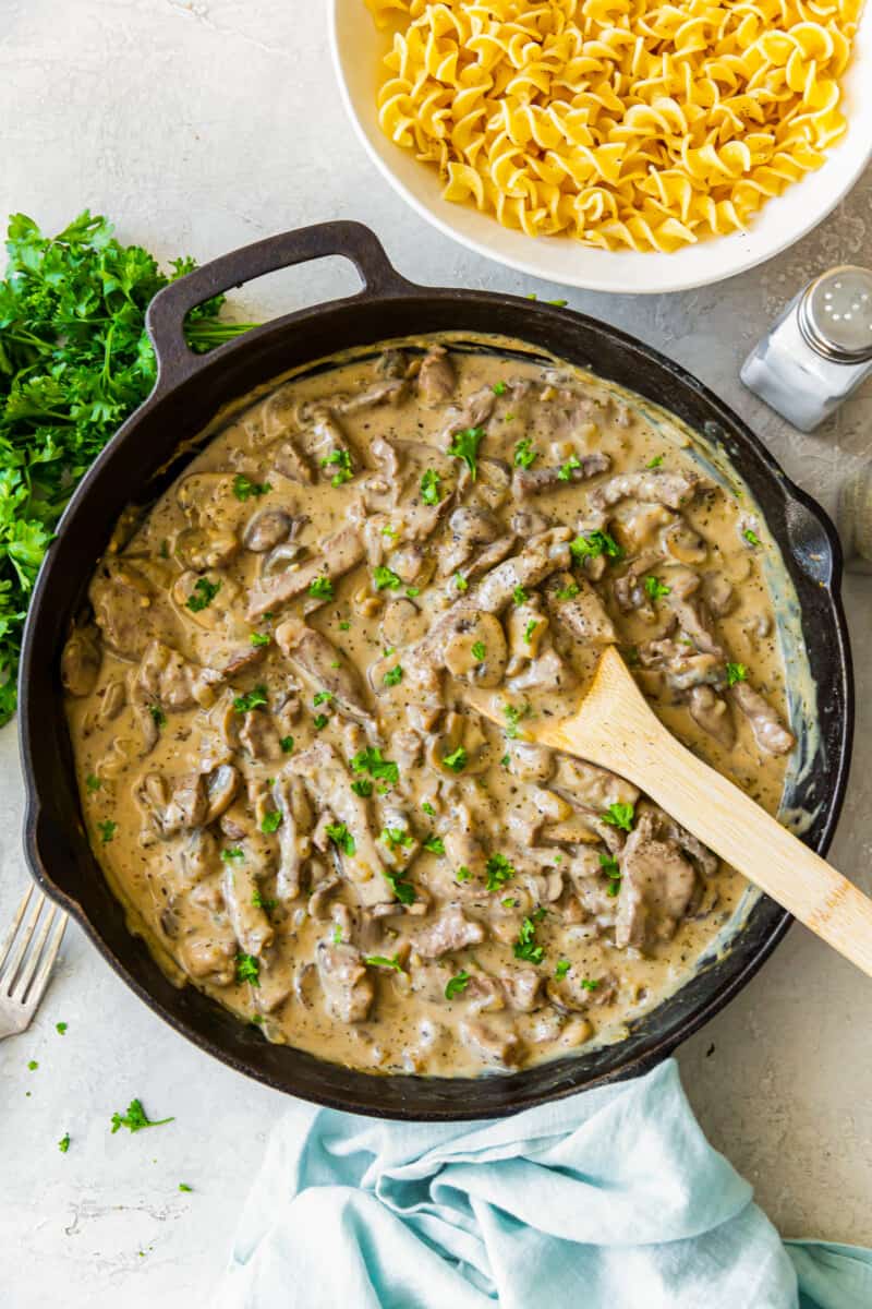 beef stroganoff sauce in a skillet with a wood spoon and cooked noodles in a white bowl