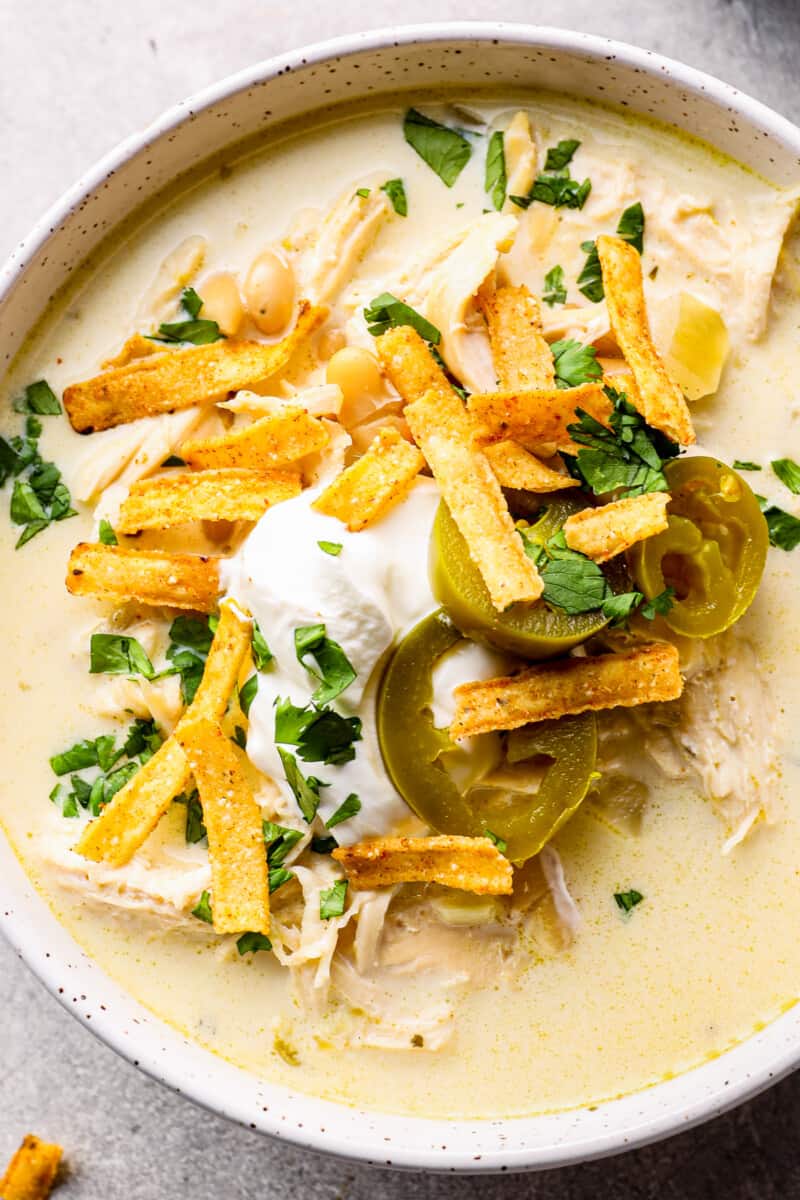 up close crockpot white chicken chili in white bowl garnished with cheese, jalapenos, and tortilla strips