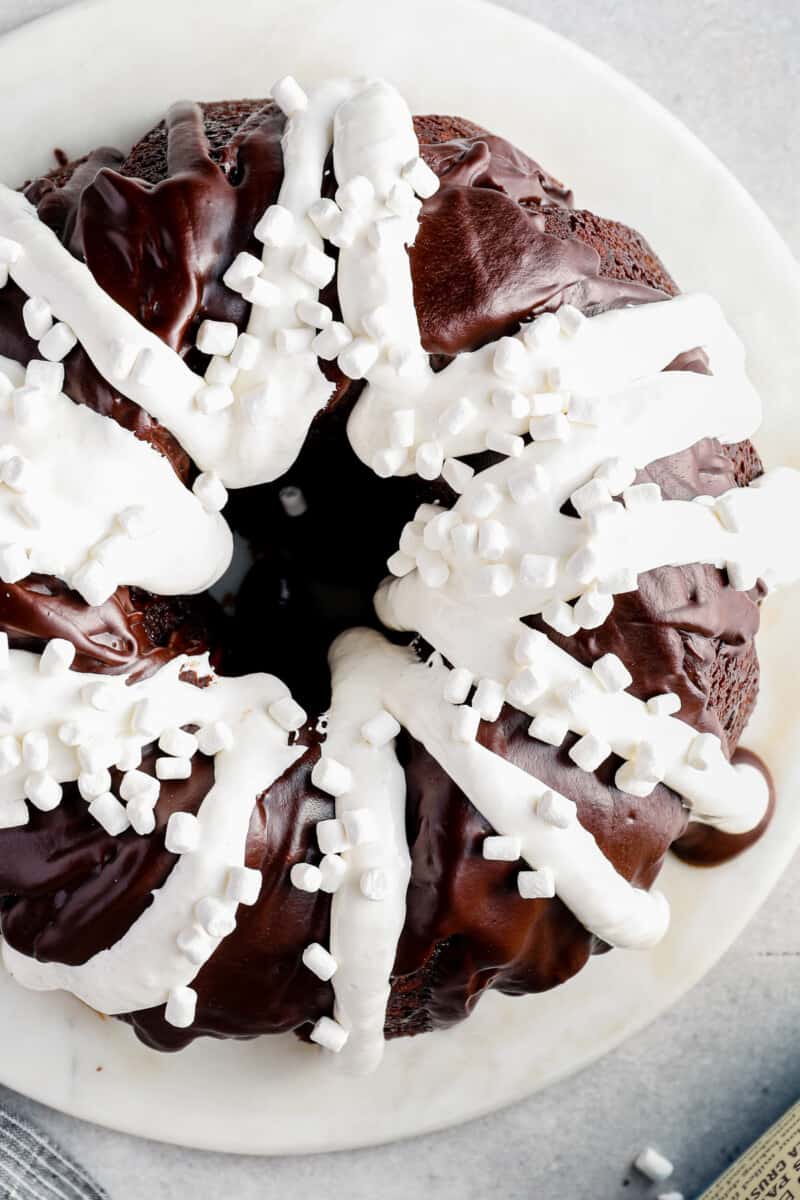 overhead image of chocolate bundt cake topped with chocolate ganache, marshmallow creme, and mini marshmallows on a white serving tray before slicing