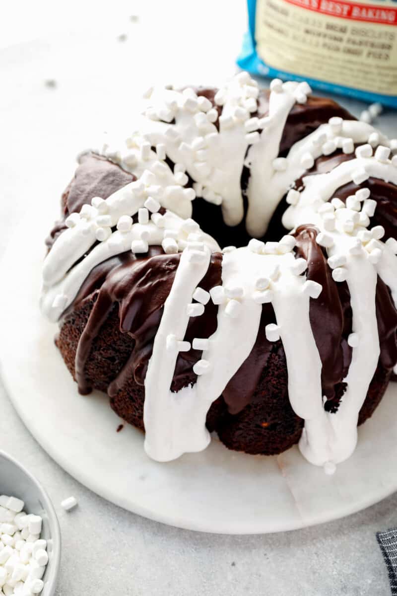 chocolate bundt cake topped with chocolate ganache, marshmallow creme, and mini marshmallows on a white serving tray before slicing