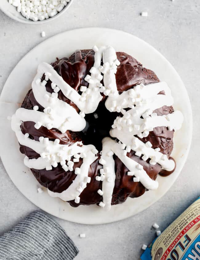 overhead image of chocolate bundt cake topped with chocolate ganache, marshmallow creme, and mini marshmallows on a white serving tray before slicing