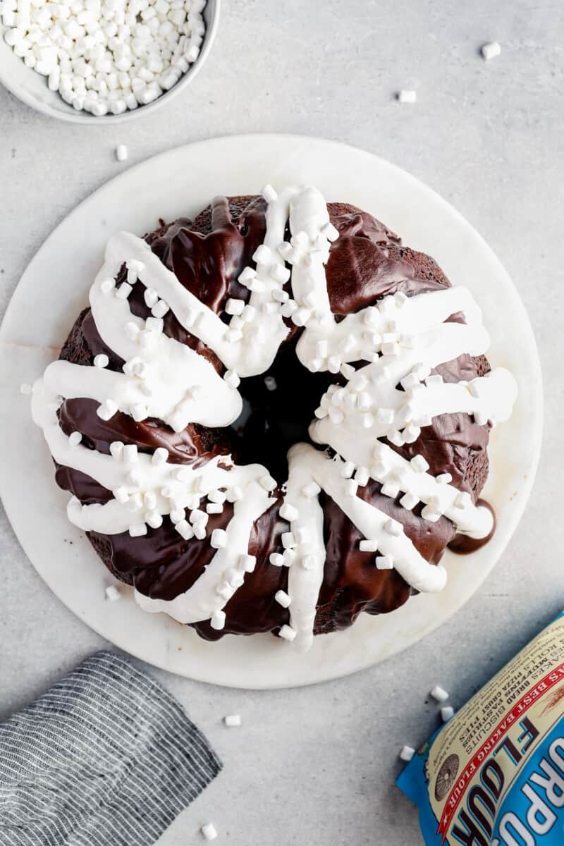 overhead image of chocolate bundt cake topped with chocolate ganache, marshmallow creme, and mini marshmallows on a white serving tray before slicing