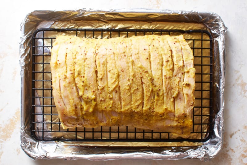 apple cider poured into the bottom of a rimmed baking sheet with a wire rack inset holding a mustard glazed raw pork tenderloin.