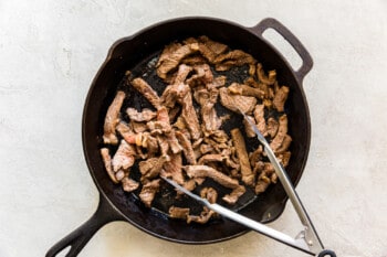 cooked strips of beef in a skillet with metal tongs