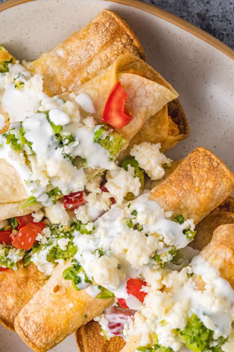 chicken taquitos on a serving plate topped with crema, guacamole, and diced tomatoes