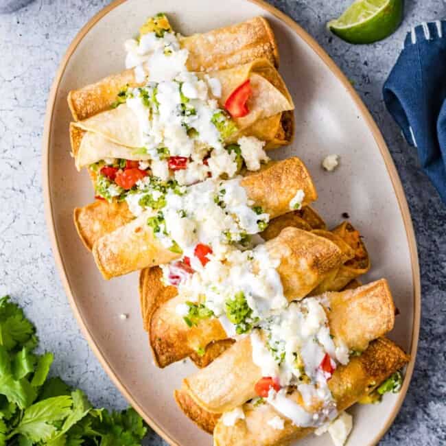 overhead image of chicken taquitos on a serving plate topped with crema, guacamole, and diced tomatoes