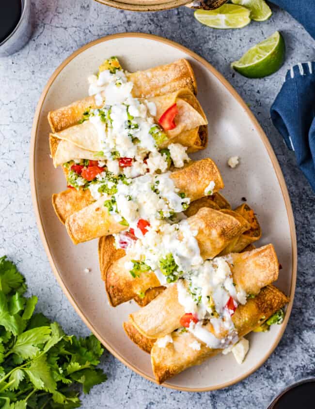 overhead image of chicken taquitos on a serving plate topped with crema, guacamole, and diced tomatoes