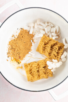 2 ramen noodle bricks in a white soup pot with seasoning and mushrooms.