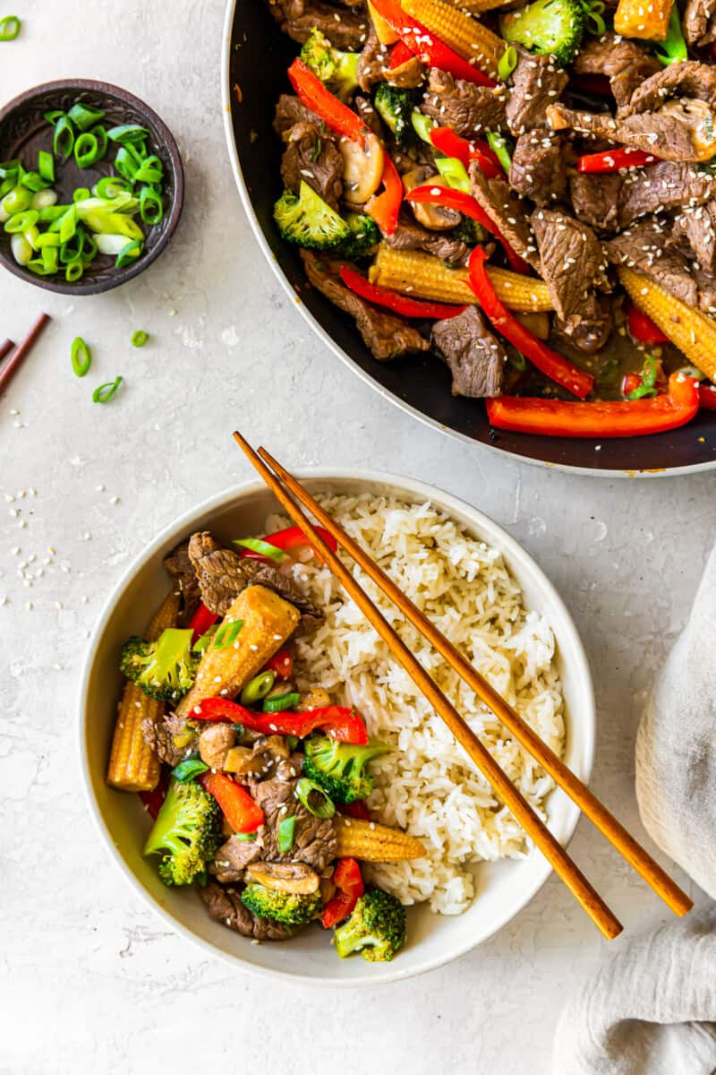 beef stir fry in a wok with a wood spoon and a serving of beef stir fry with rice in a white bowl with chopsticks