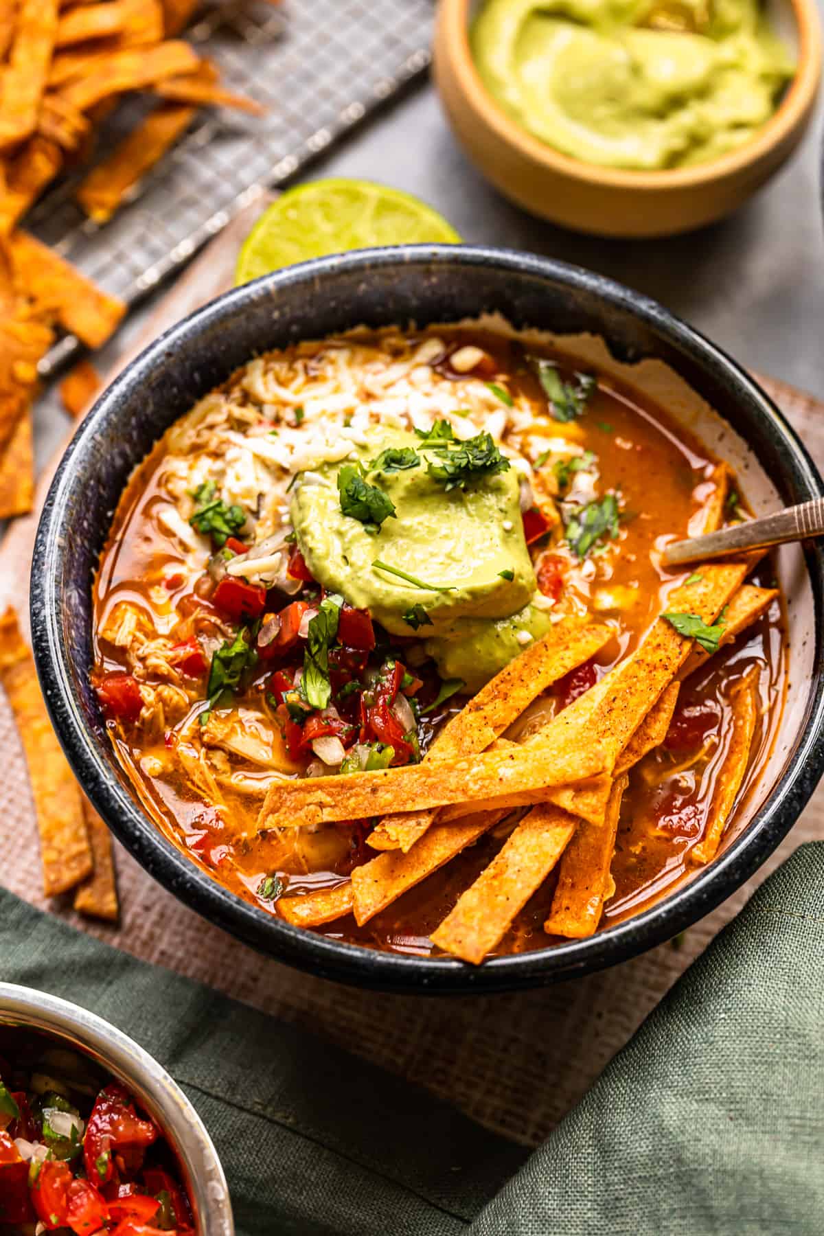 chicken fajita soup in a black bowl topped with cheese, guacamole, diced tomatoes, and tortilla strips with a spoon