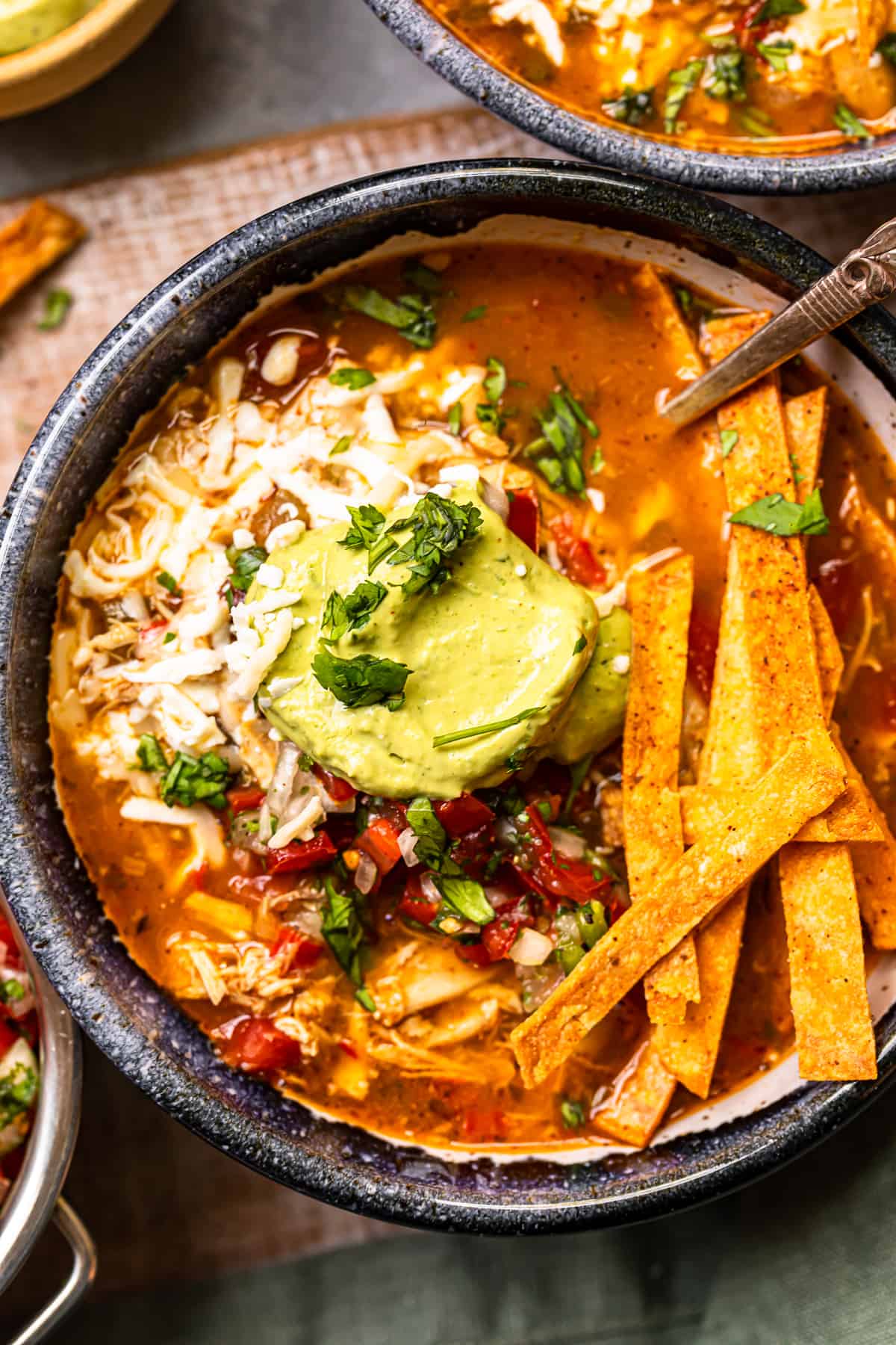 close up image of chicken fajita soup in a black bowl topped with cheese, guacamole, diced tomatoes, and tortilla strips with a spoon