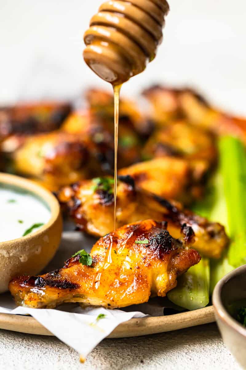 using a honey dipper to drizzle honey onto hot honey chicken wings on a serving platter