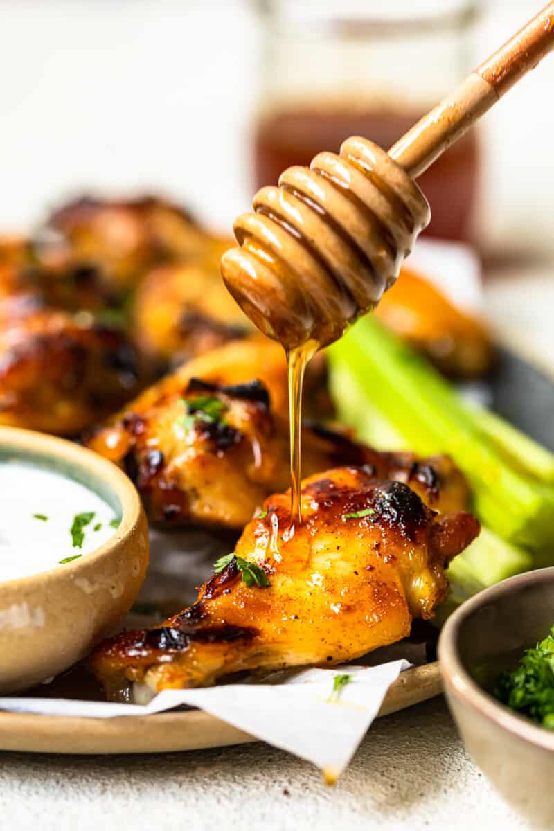 using a honey dipper to drizzle honey onto hot honey chicken wings on a serving platter