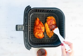 hand brushing bbq sauce onto 2 chicken breasts in air fryer basket