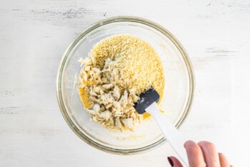 hand using a spatula to fold crab meat and bread crumbs into spices in a glass bowl