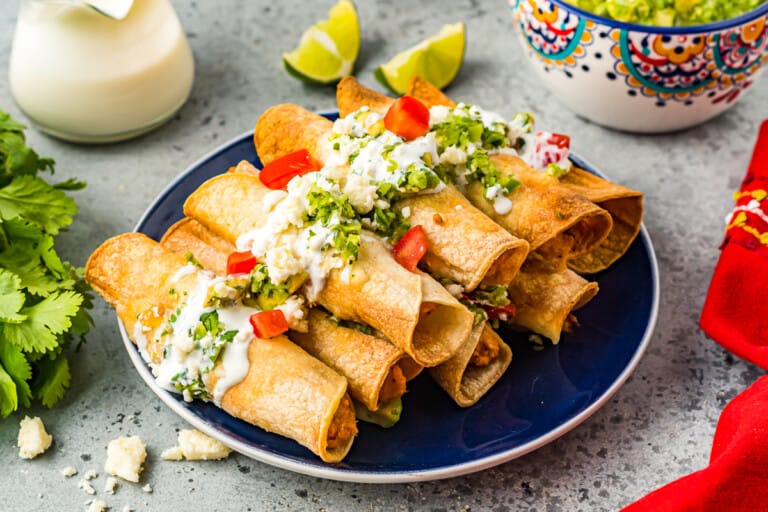 A blue plate holds a stack of golden, crispy taquitos topped with diced tomatoes, guacamole, and crumbled cheese sauce. Fresh cilantro, lime wedges, a colorful bowl of guacamole, and a red napkin are arranged around the plate on a gray surface.