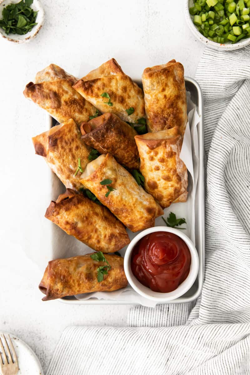 overhead image of cheeseburger egg rolls on a serving tray with a side of ketchup in a white bowl