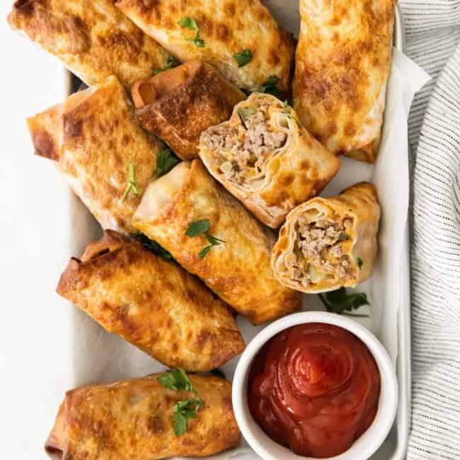 overhead image of cheeseburger egg rolls on a serving tray with a side of ketchup in a white bowl