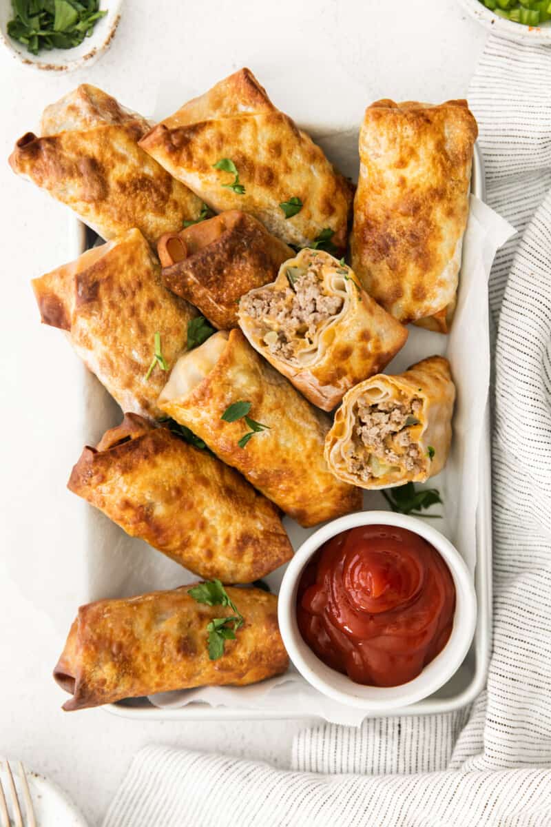 overhead image of cheeseburger egg rolls on a serving tray with a side of ketchup in a white bowl