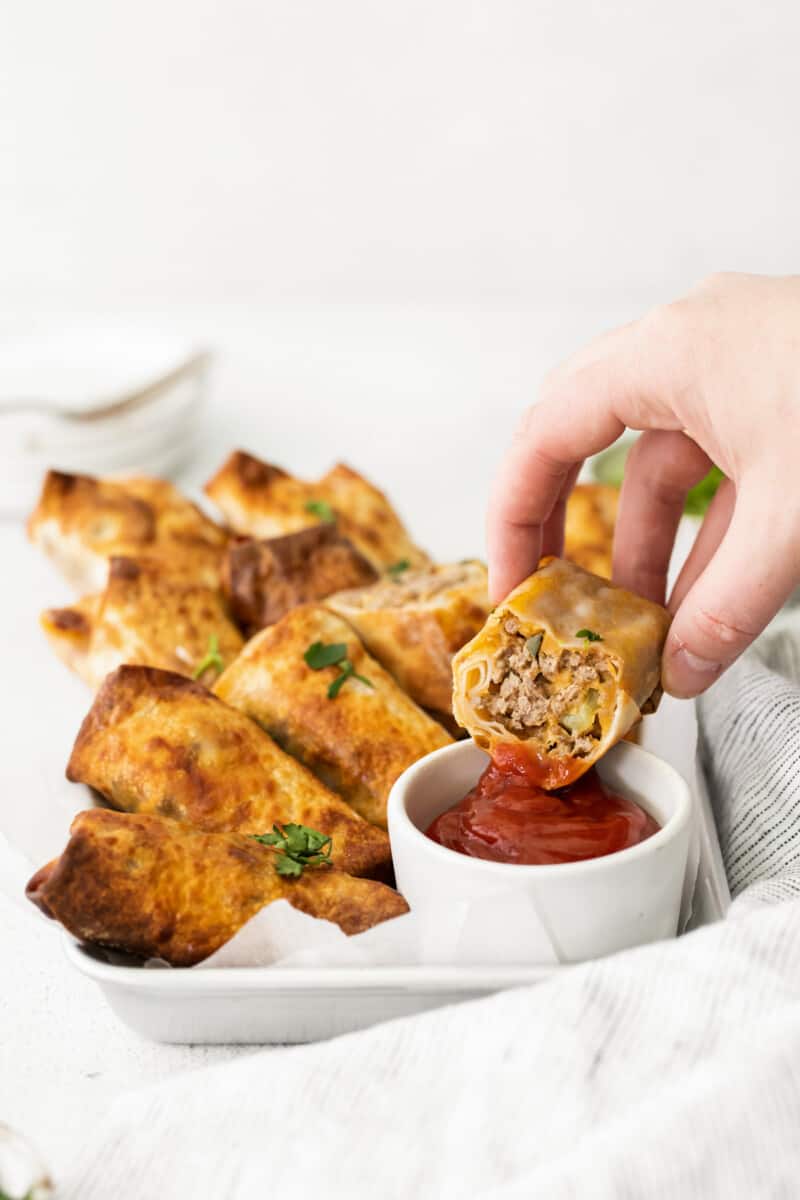 hand dipping cheeseburger egg roll into ketchup in a white bowl