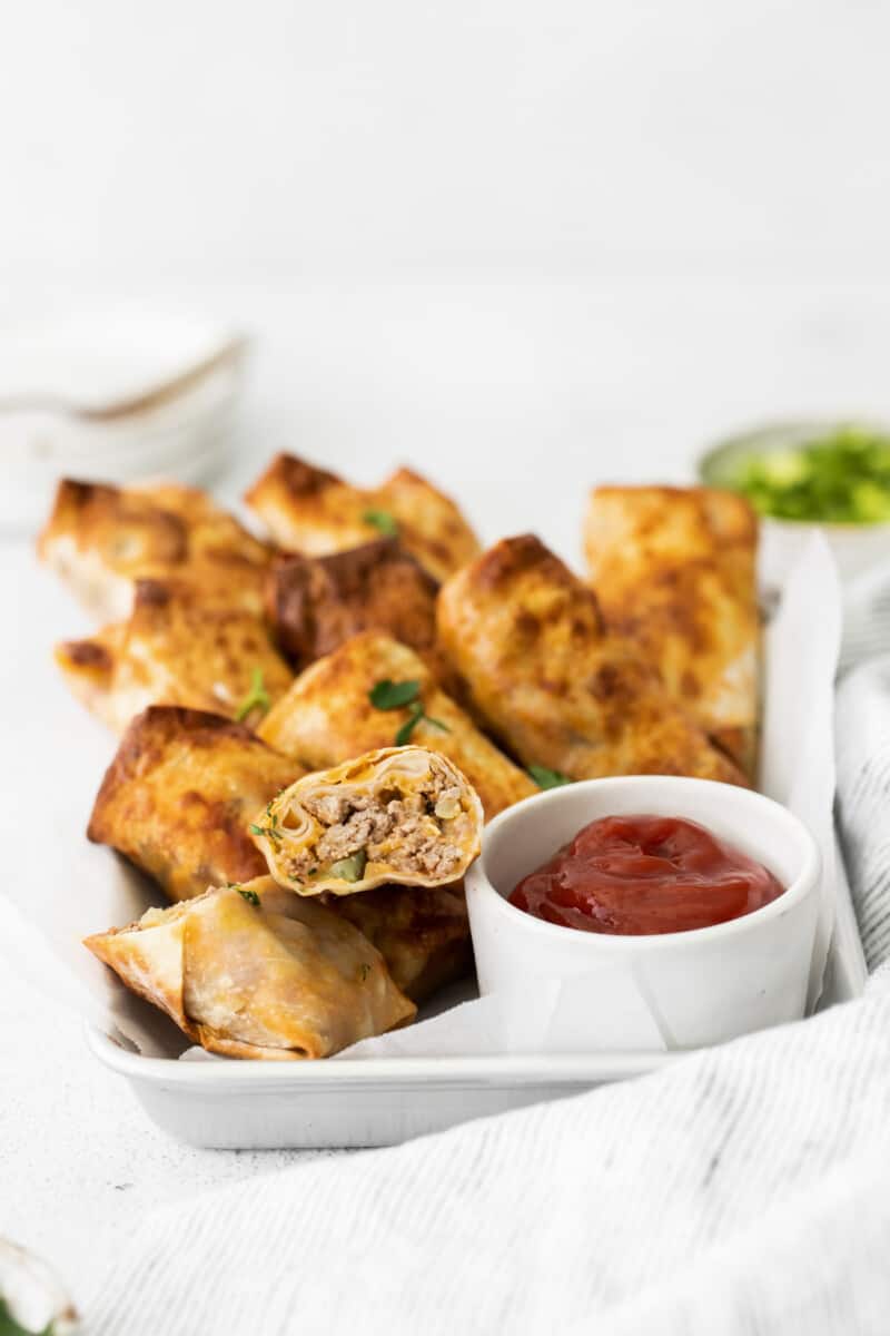 cheeseburger egg rolls with one cut in half showing the filling on a serving tray with a side of ketchup in a white bowl